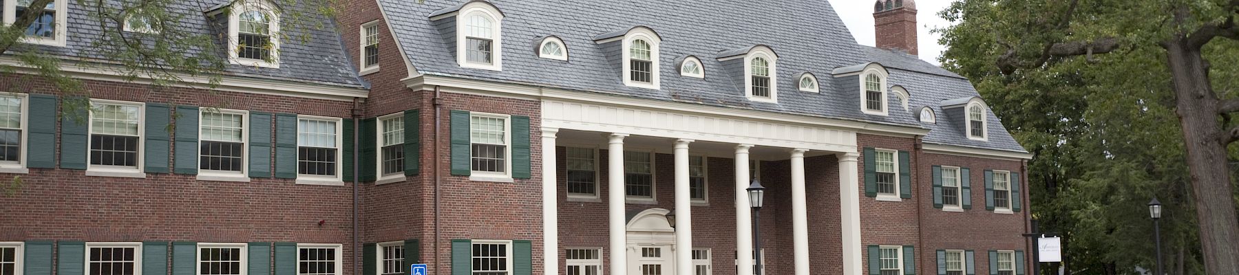 A brick collegiate-style building with a central portico, dormer windows, and a manicured entrance, set among trees and a paved lot.