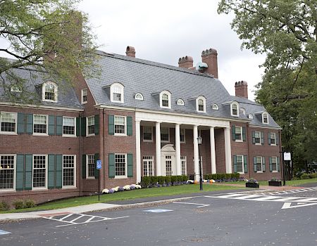 A brick collegiate-style building with a central portico, dormer windows, and a manicured entrance, set among trees and a paved lot.