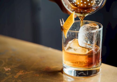 A whiskey glass with ice and an orange twist, being poured from a shaker, on a wooden bar top, with warm bar lighting. Ending with a period.