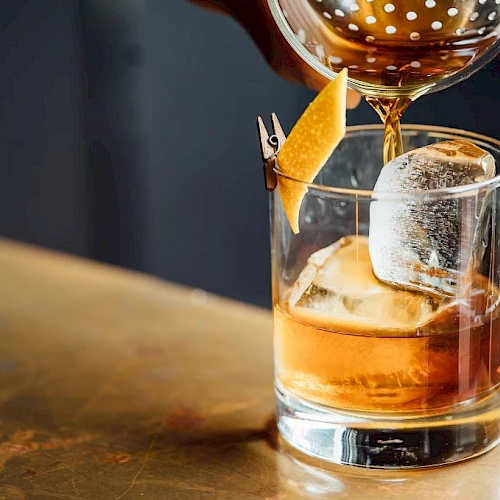 A whiskey glass with ice and an orange twist, being poured from a shaker, on a wooden bar top, with warm bar lighting. Ending with a period.