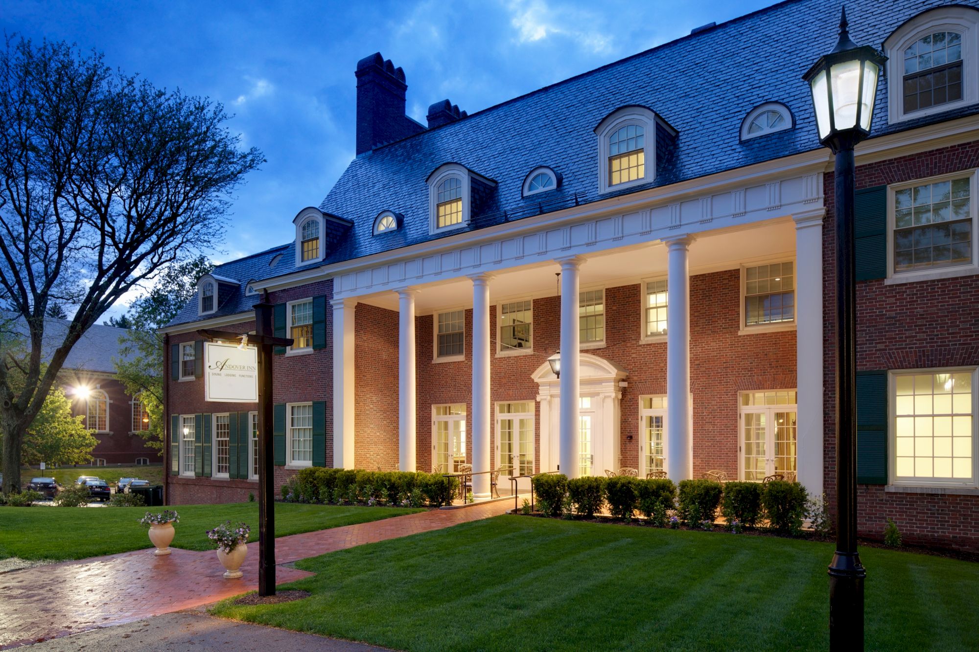 The image shows a large brick building with white columns, a slate roof, and a well-lit entrance, set against a twilight sky.