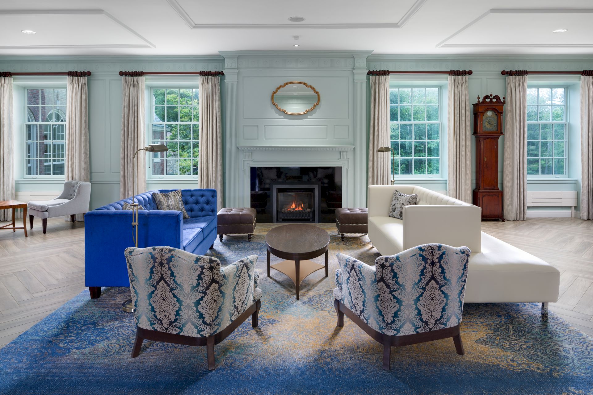 A modern living room with a fireplace, blue and white sofas, patterned chairs, a blue rug, grandfather clock, and large windows.