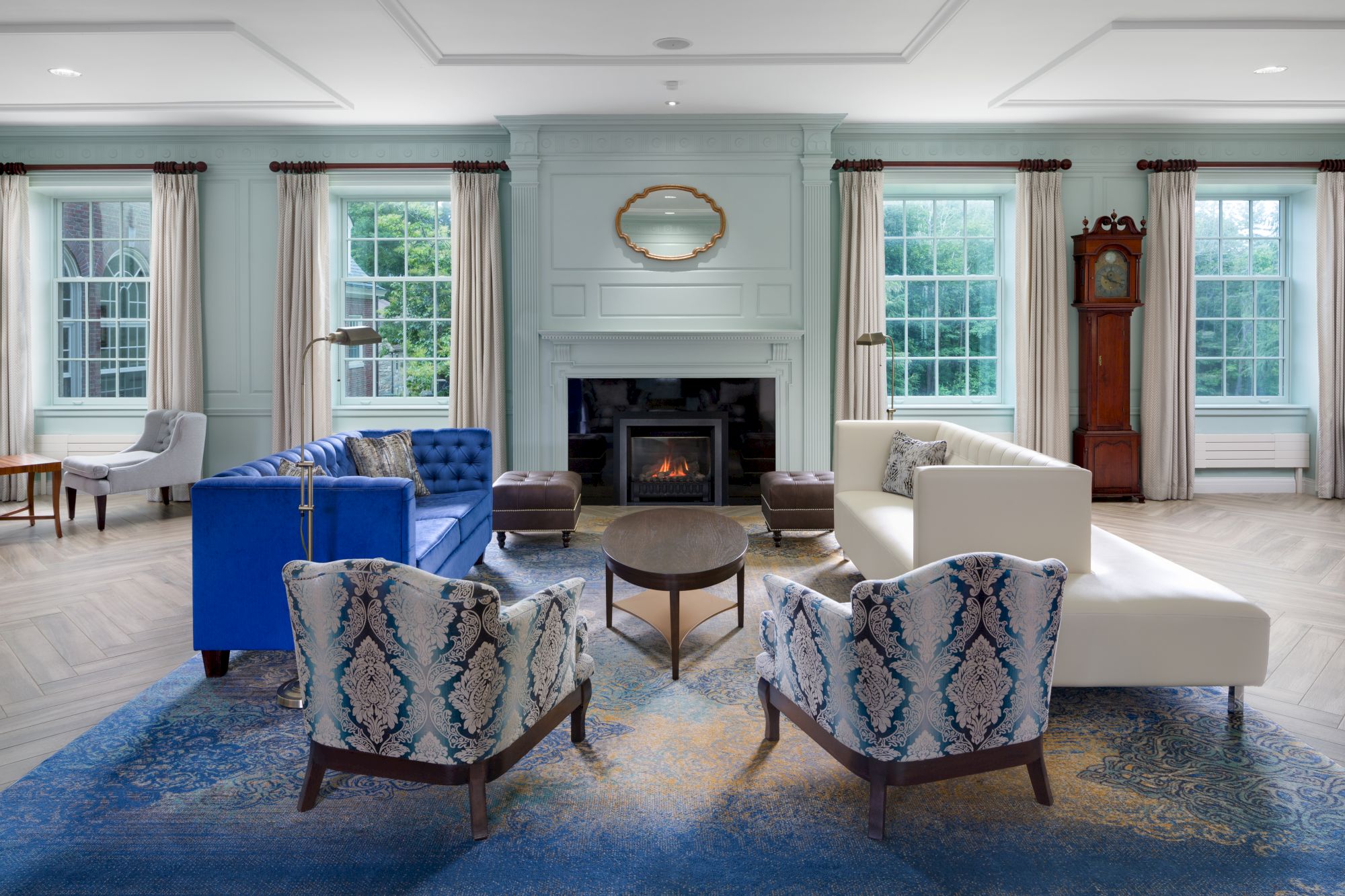 A modern living room with a fireplace, blue and white sofas, patterned chairs, a blue rug, grandfather clock, and large windows.