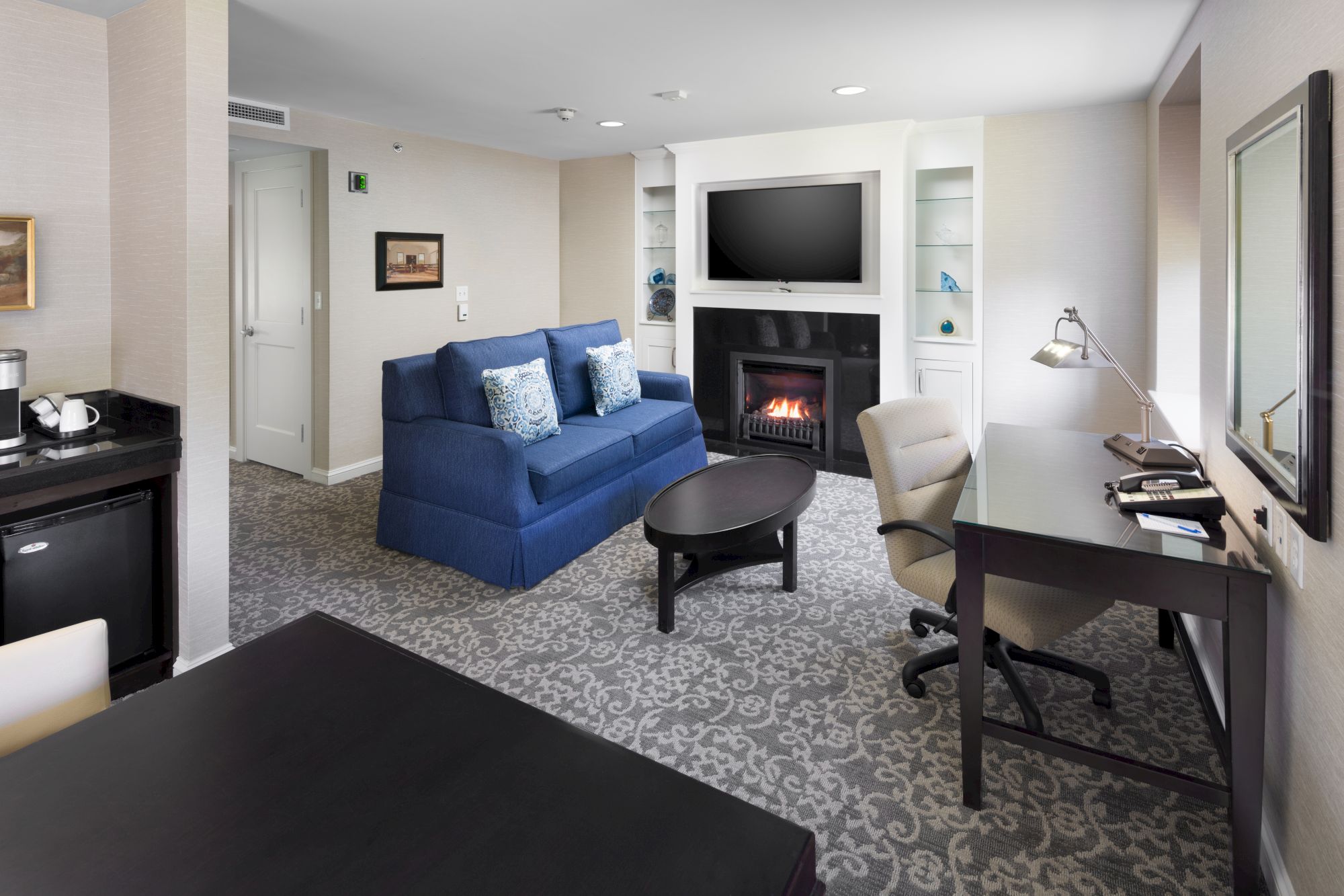 The image shows a modern living area with a blue sofa, TV above a fireplace, desk, chair, and patterned carpet, creating a cozy ambiance.