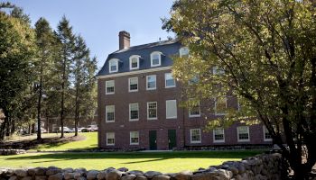 A brick building with multiple windows, surrounded by trees and a stone wall, under a clear blue sky.