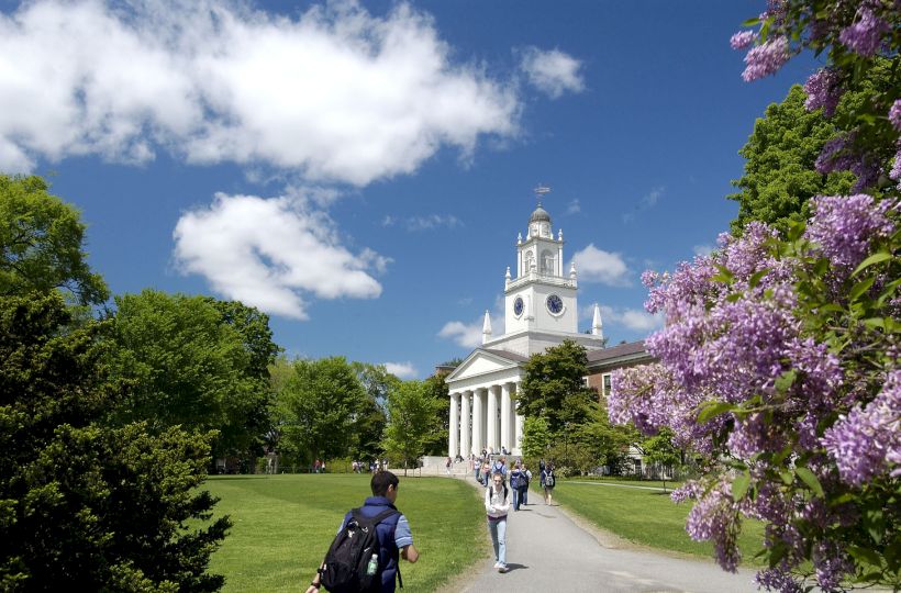 A scenic campus with a prominent building featuring a clock tower, surrounded by trees and flowers. People walk along the path.