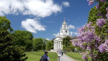 A group of people walk towards a building with columns and a clock tower, surrounded by trees and blooming flowers under a blue sky.