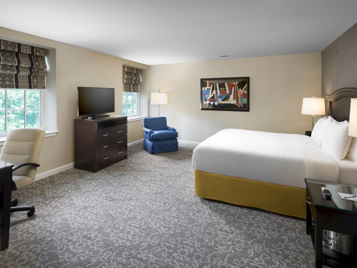 A hotel room with a large bed, TV, seating area, desk, and window. The decor is modern with tasteful art on the wall.