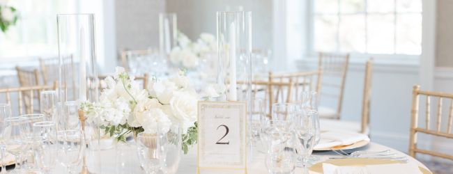 A beautifully set round table with elegant glassware, a white floral centerpiece, gold accents, and a table number "2" in a bright room.