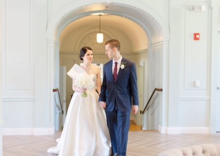 A couple is walking indoors; the bride holds a bouquet and wears a gown, while the groom is in a suit, both looking at each other.