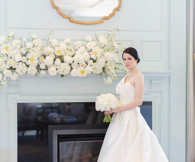 A bride in a white gown stands by a fireplace adorned with flowers, holding a bouquet, with a mirror above.