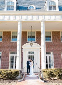 A couple stands at the entrance of a grand brick building with tall columns and multiple windows, under a sunny sky.