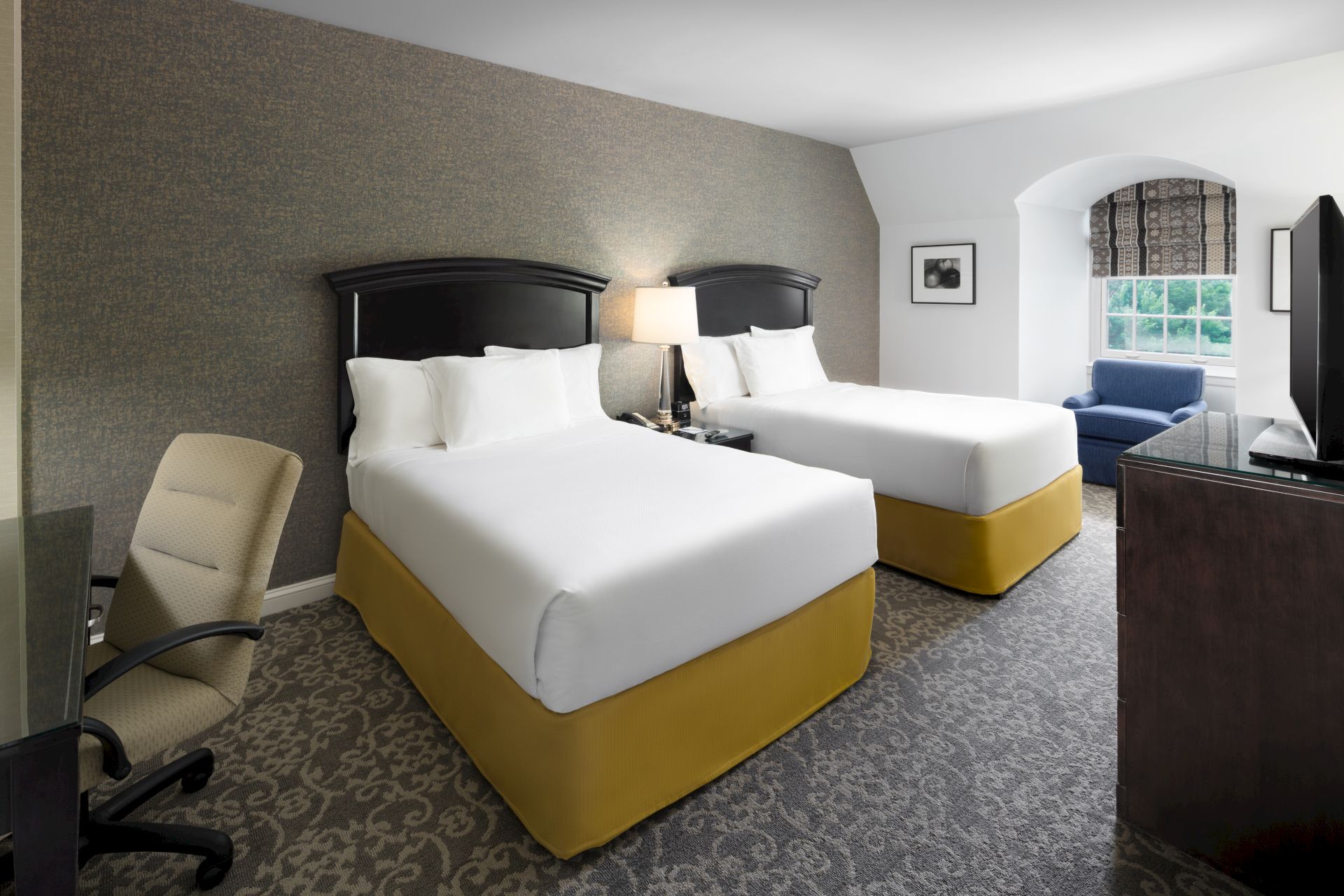 A hotel room with two beds, a desk and chair, a lamp, a window, and a TV. The beds have white linens and yellow bases.