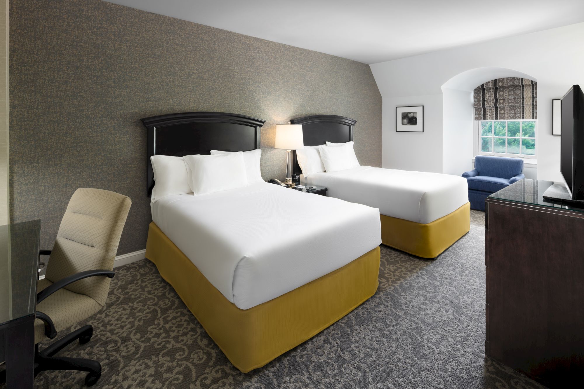 A hotel room with two beds, a desk and chair, a lamp, a window, and a TV. The beds have white linens and yellow bases.