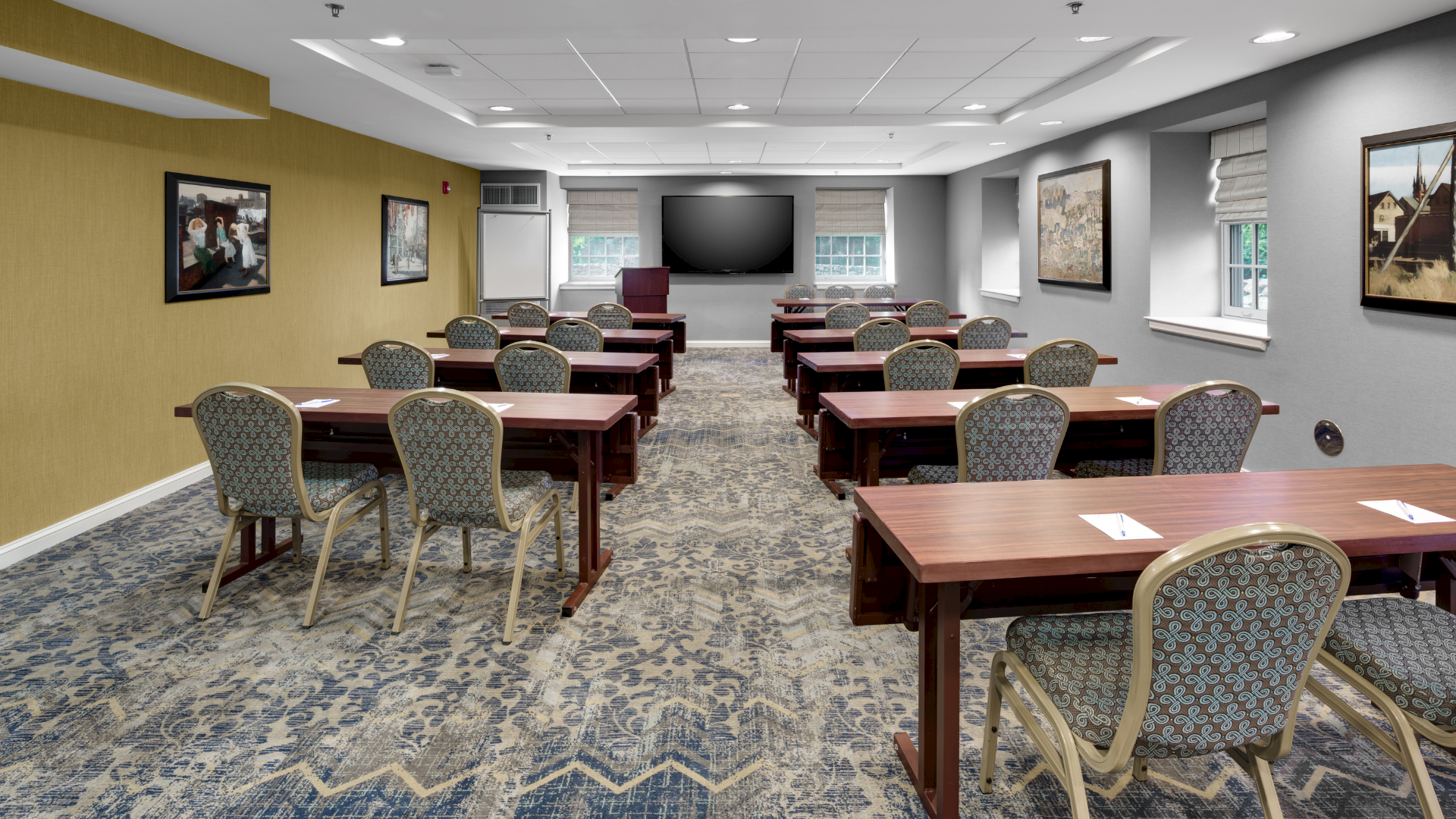 A bright conference room with rows of wooden tables, patterned chairs, wall art, and a large flat-screen TV.