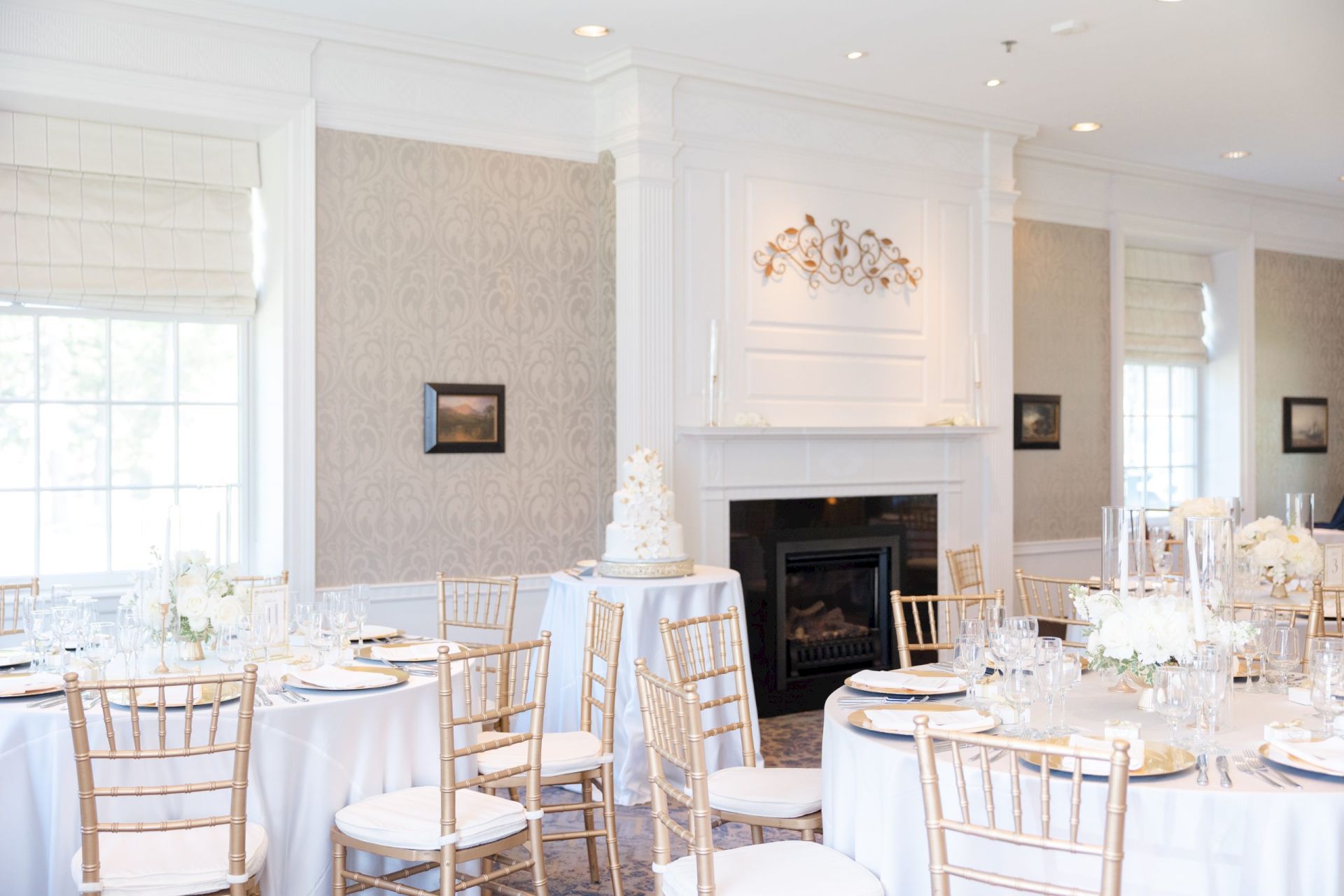 Elegant wedding reception room with round tables, gold chairs, white cake, and a fireplace in the background.