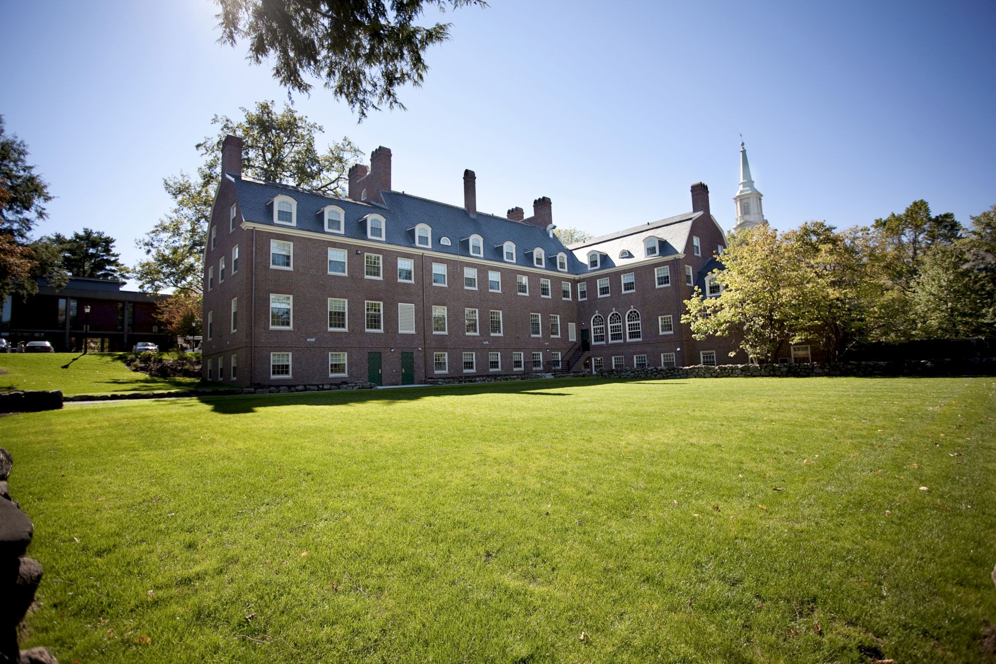 A red-brick building with multiple windows and chimneys, set beside a spacious green lawn under a clear blue sky on a sunny day.