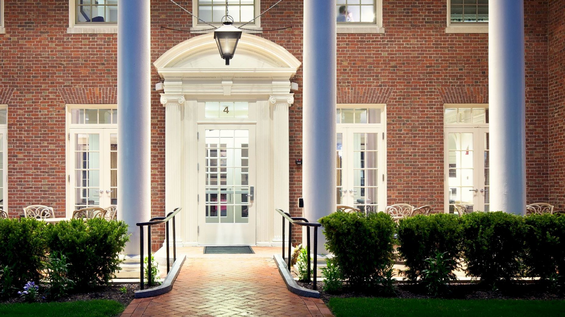 The image shows the exterior of a building with brick walls, tall columns, and a lit entrance surrounded by windows and greenery.