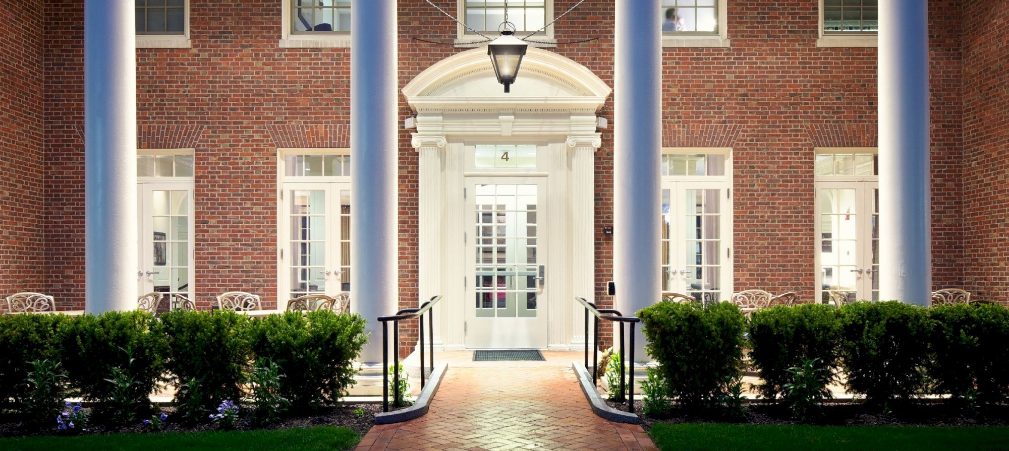 The image shows the exterior of a building with brick walls, tall columns, and a lit entrance surrounded by windows and greenery.