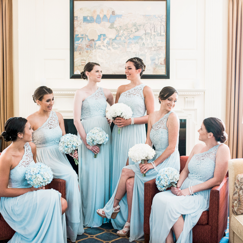 A group of women in matching light blue dresses, each holding a white bouquet, sitting and standing in a well-lit room.