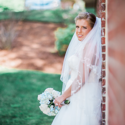 A bride in a white dress stands by a brick wall, holding a bouquet of white flowers, with a garden in the background.