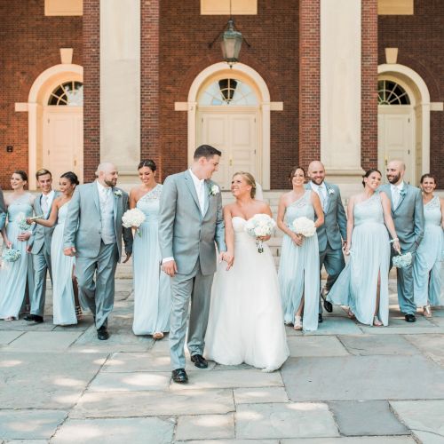 A wedding party stands in front of a building, with the couple in the center and the bridal party in matching outfits surrounding them.