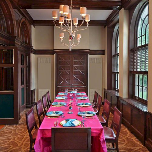 A formal dining room with a long table, red tablecloth, chairs, chandelier, and large windows creating an elegant atmosphere.
