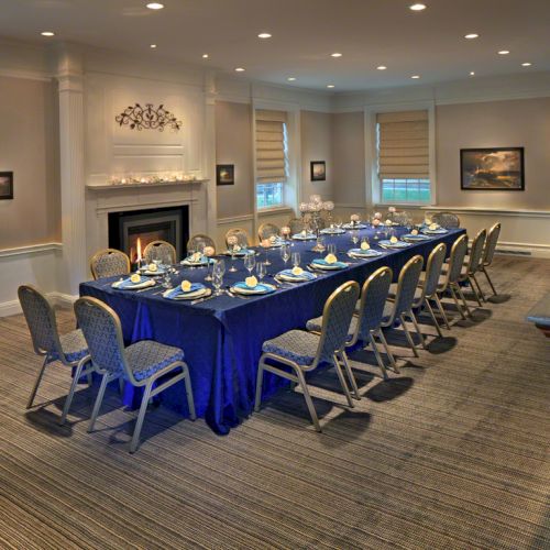 An elegant conference room with a U-shaped table set with place settings, chairs, a fireplace, windows, and framed artwork on the walls.