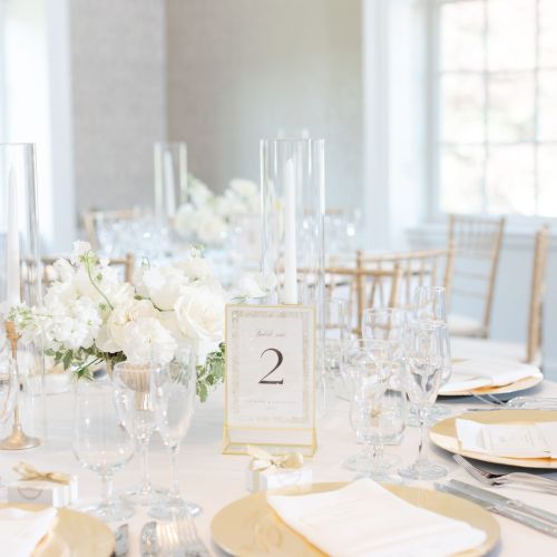 Elegant table setting with white flowers, glassware, and gold accents, marked by a table number in a bright, airy room.