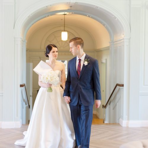 A bride and groom holding hands, walking indoors in a light-filled setting. The bride holds a bouquet; both look at each other warmly.