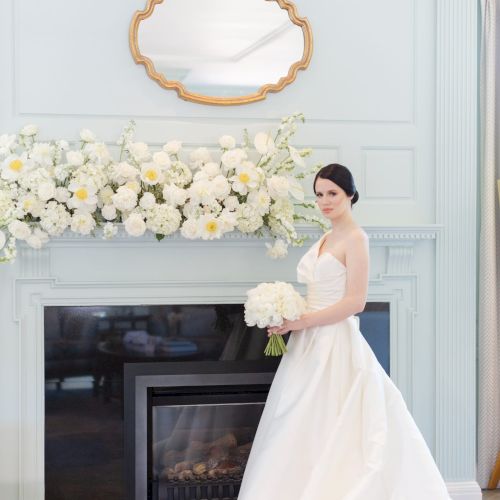 A bride in a white gown stands by a fireplace decorated with white flowers, beneath an ornate mirror, holding a bouquet.