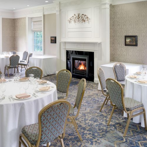 The image shows a bright banquet room with round tables set for an event, a fireplace, large windows, and elegant décor.