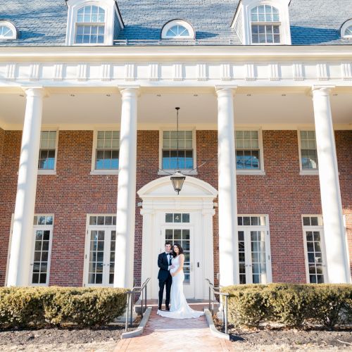 A couple is standing in front of a large brick building with tall white columns and multiple windows, under a sunny sky.