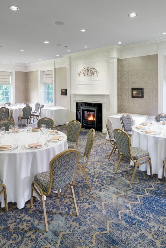 The image shows an elegant dining room set up for an event with round tables, chairs, a fireplace, and a large TV on the wall.