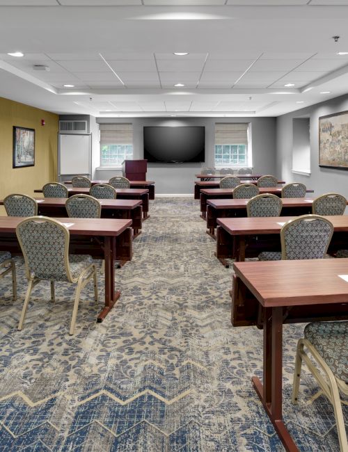 The image shows a conference room with rows of tables and chairs facing a large screen, decorated with framed pictures on the walls.