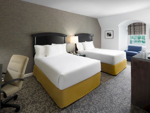 The image shows a hotel room with two beds, a desk with a chair, a lamp, a TV, a window, and a blue chair by the corner wall.