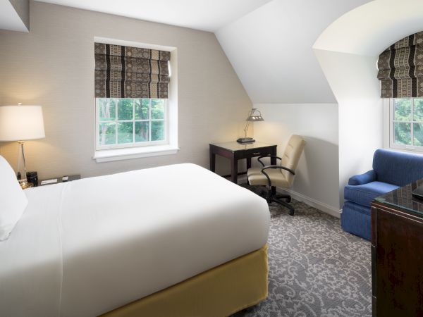 A cozy hotel room features a bed, desk with a lamp, chair, window with blinds, and an armchair, perfect for relaxing and work.