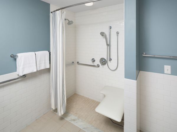 An accessible shower with grab bars, a foldable seat, a handheld showerhead, and a curtain. Walls are tiled and painted blue.