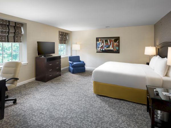 A spacious hotel room with a king bed, two nightstands, a dresser with a TV, a blue chair, a desk by the window, and soft lighting.