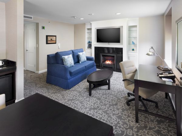 A modern living room with a blue sofa, flat-screen TV, fireplace, desk, and chairs, featuring a patterned carpet and subtle decor.