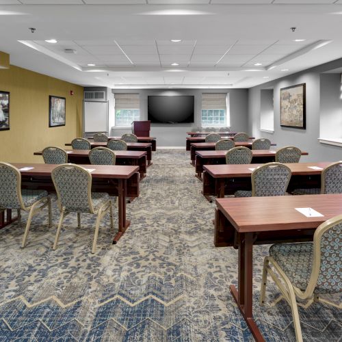 The image shows an empty conference room with rows of chairs and tables, a large screen at the front, and artwork on the walls.
