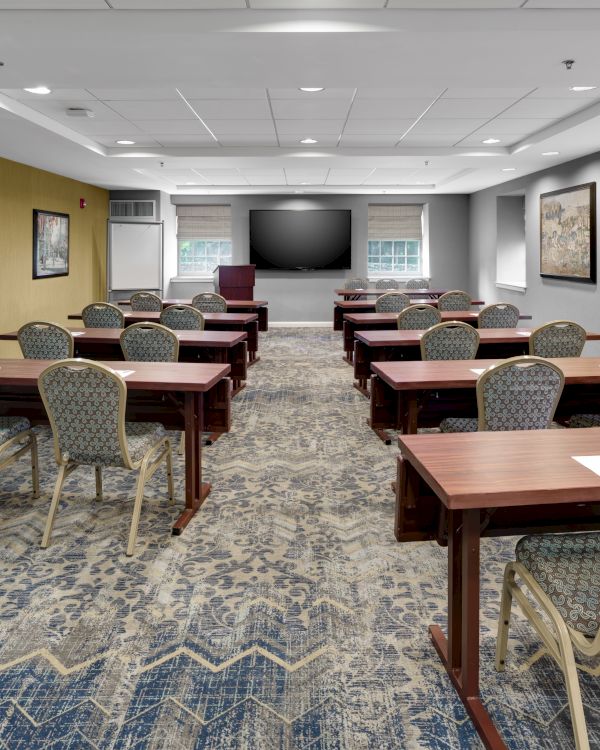The image shows an empty conference room with rows of chairs and tables, a large screen at the front, and artwork on the walls.