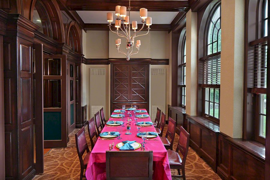 Elegant dining room with a long table, pink tablecloth, blue plates, wooden chairs, chandelier, and large arched windows.