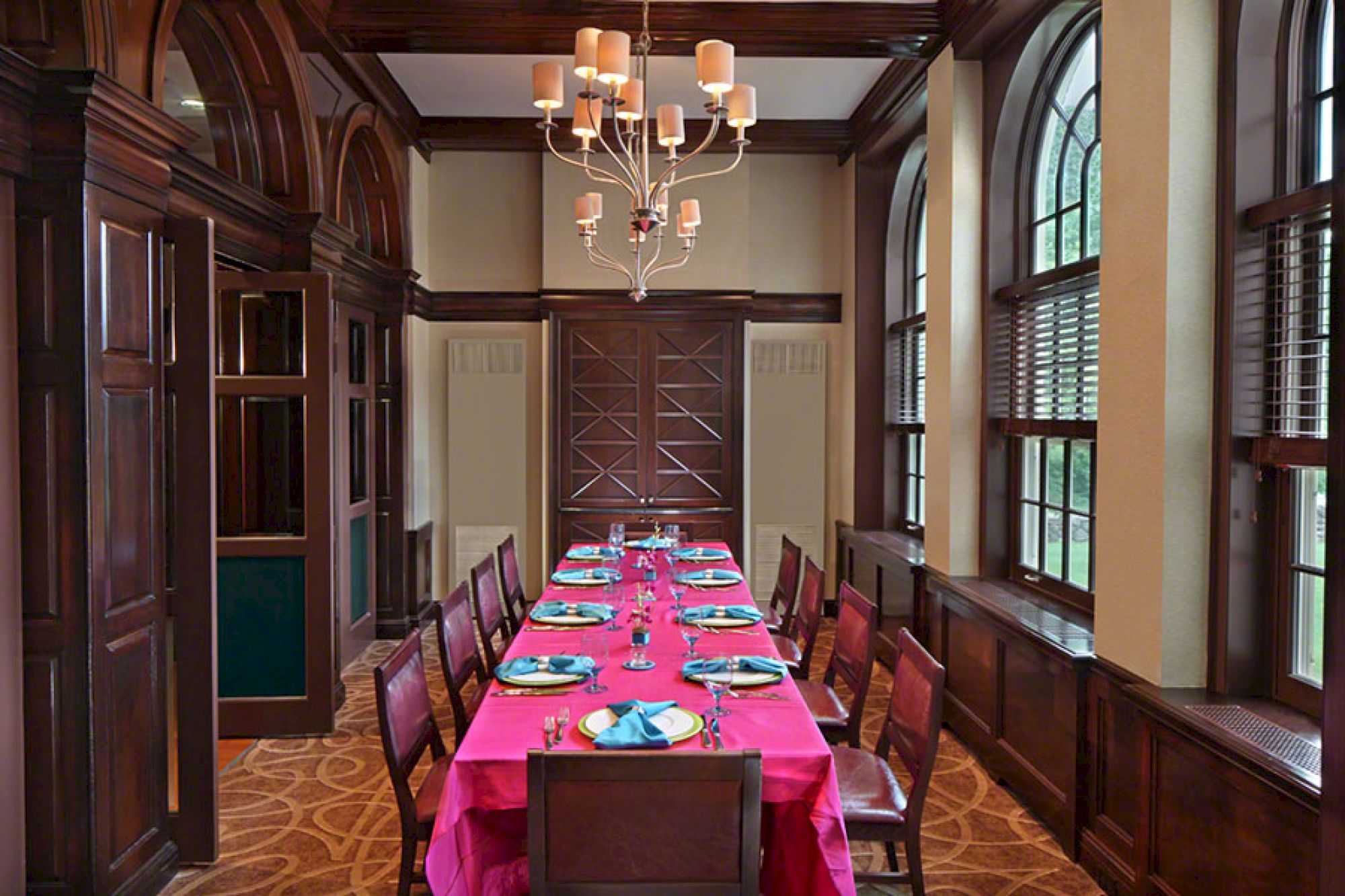 Elegant dining room with a long table, pink tablecloth, blue plates, wooden chairs, chandelier, and large arched windows.