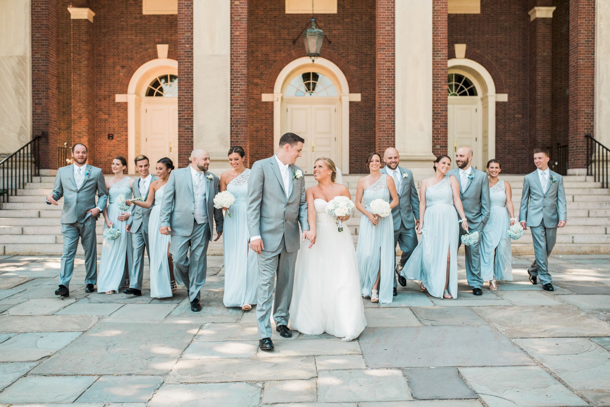 A wedding party poses outdoors in front of a building with arches, with the couple in the center and bridesmaids and groomsmen surrounding them.