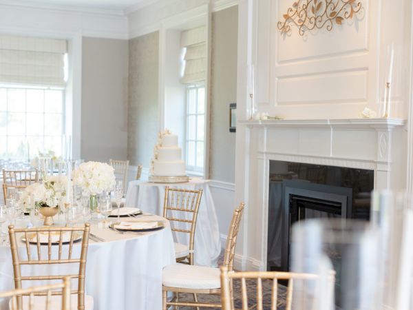 Elegant dining room set for an event, with white tablecloths, gold chairs, floral centerpieces, and a decorative mantle.