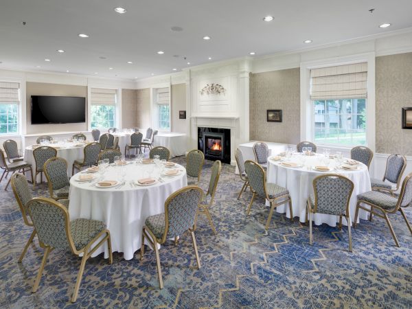 The image shows a banquet hall set up with round tables, chairs, a fireplace, and a TV. The room has windows and patterned carpet.