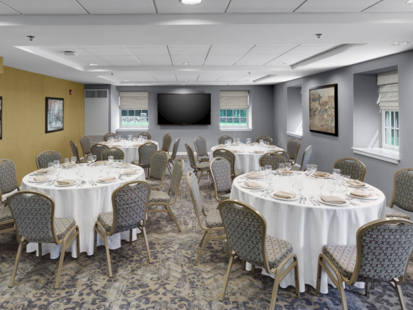 A banquet room with round tables, white tablecloths, beige chairs, framed pictures on gray and yellow walls, a large screen at the far wall, and windows letting in light.