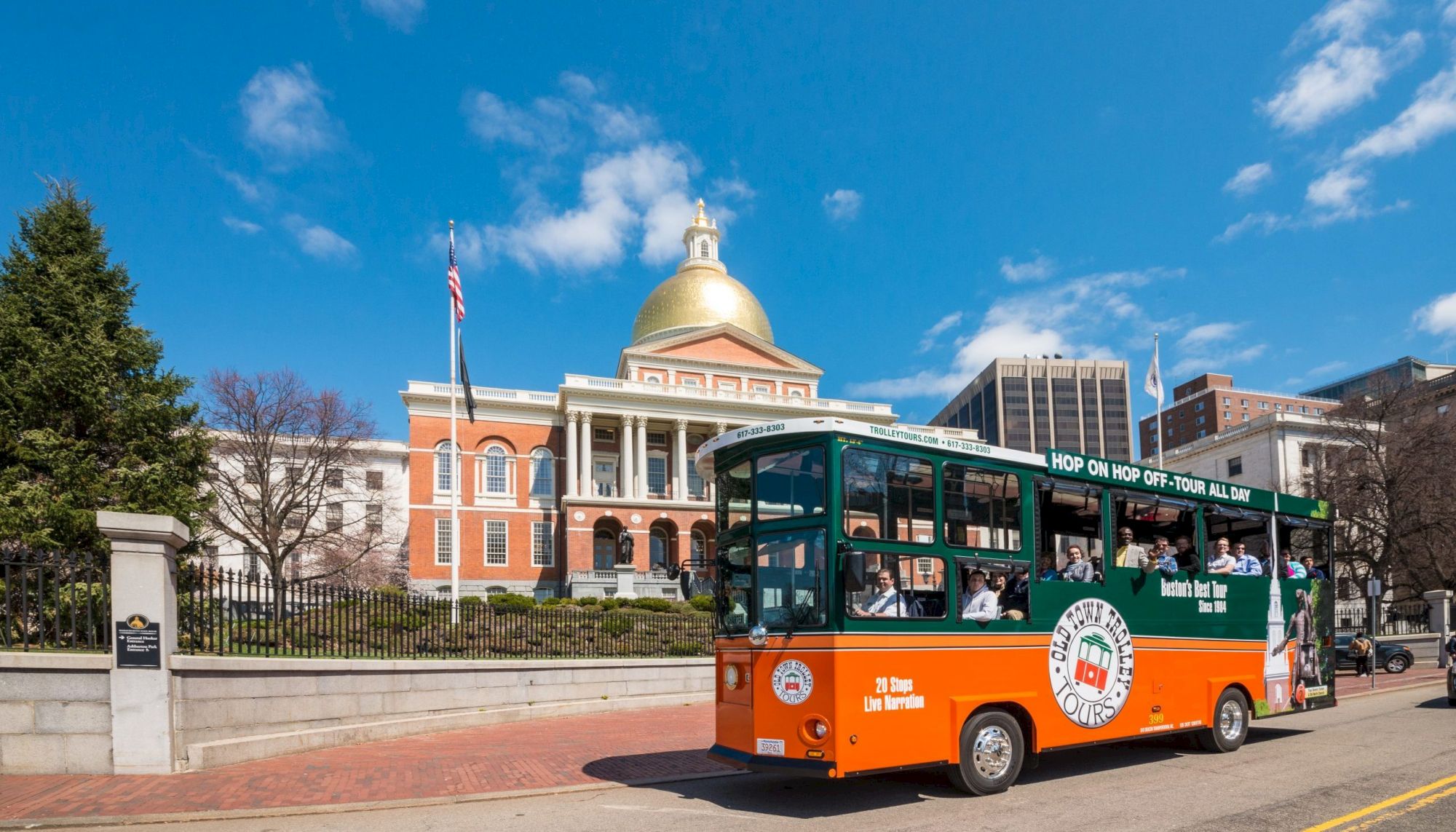 A trolley bus drives in front of a historic building with a gold dome and red brick facade under a clear blue sky.