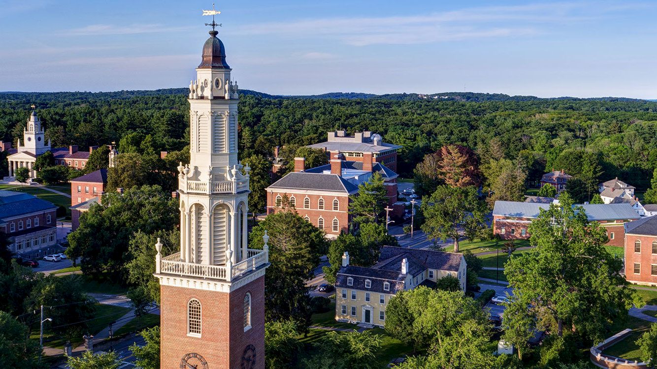 An aerial view of a historic campus with a prominent clock tower, surrounded by lush greenery and multiple brick buildings.